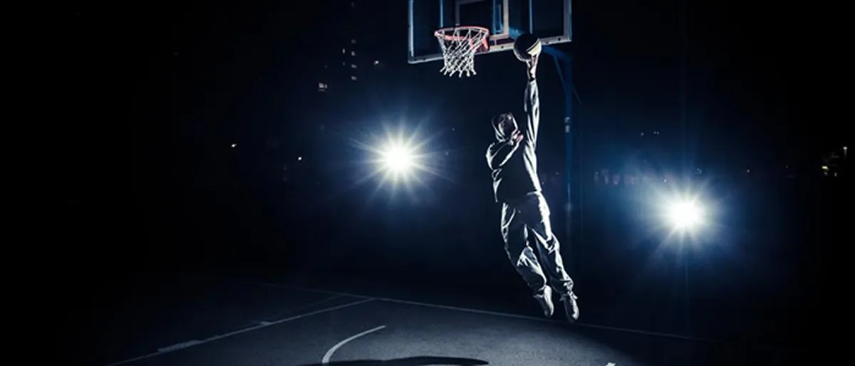 A basketball player silhouetted mid-air against floodlights, laying the ball into the hoop on a dark outdoor court