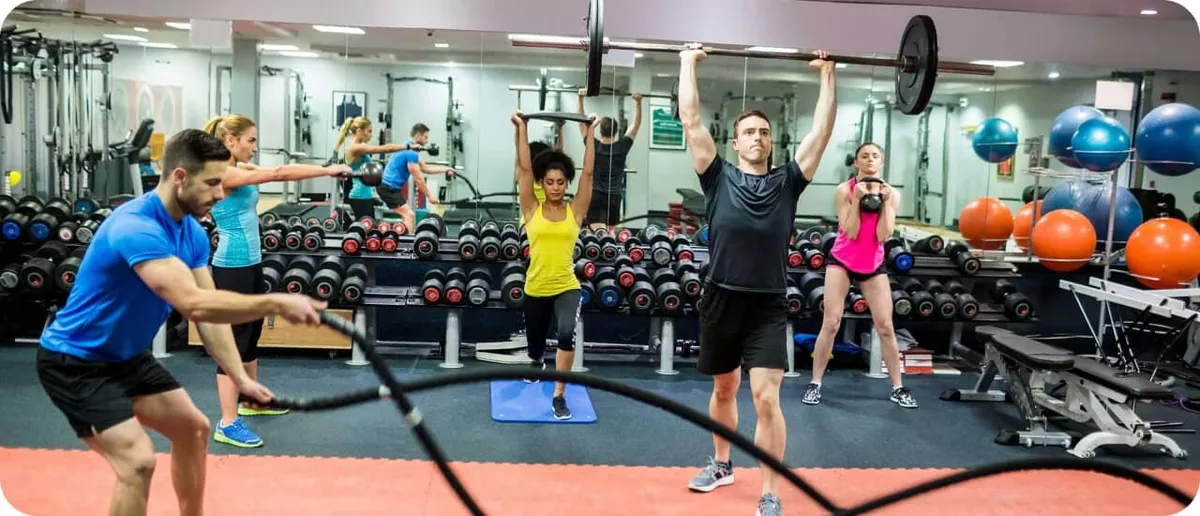 A busy gym floor with people using battle ropes, pressing a barbell overhead, swinging a kettlebell and lifting dumbbells