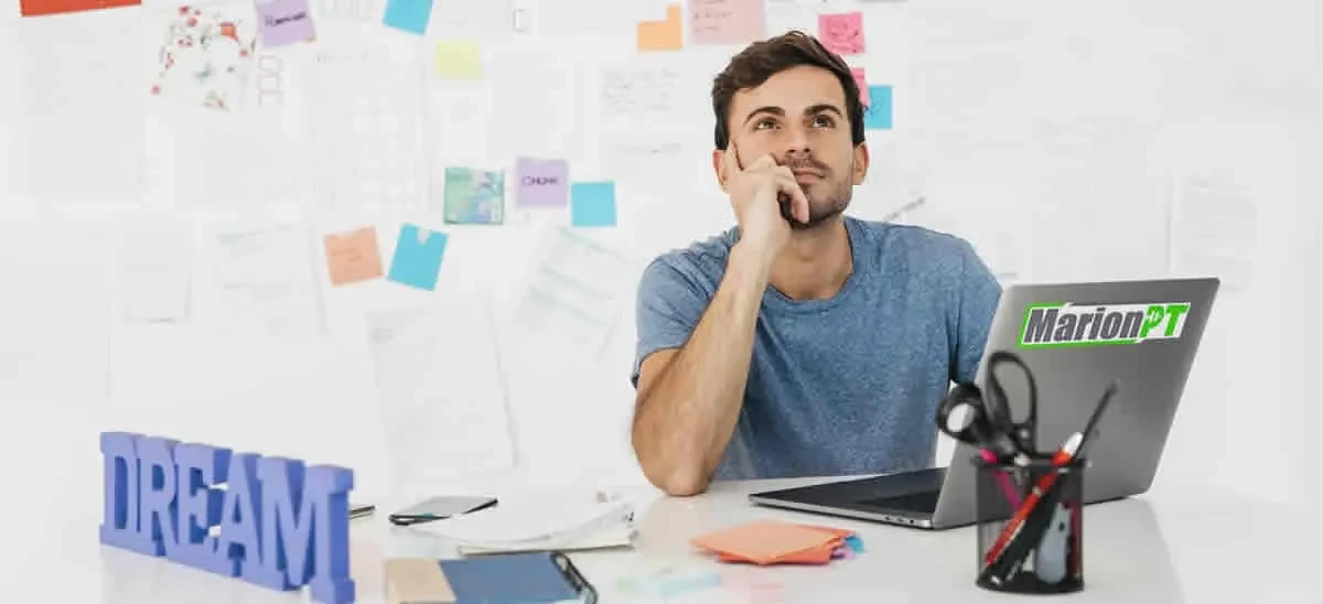 A man daydreaming at a desk covered in sticky notes and paperwork, beside a laptop and a sign reading DREAM