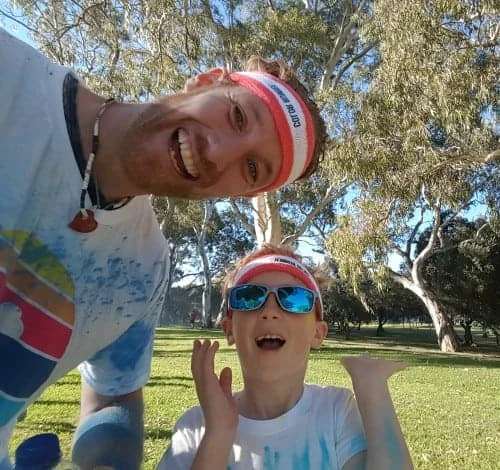 Henk and his son grinning at the camera in matching headbands at a park fun run