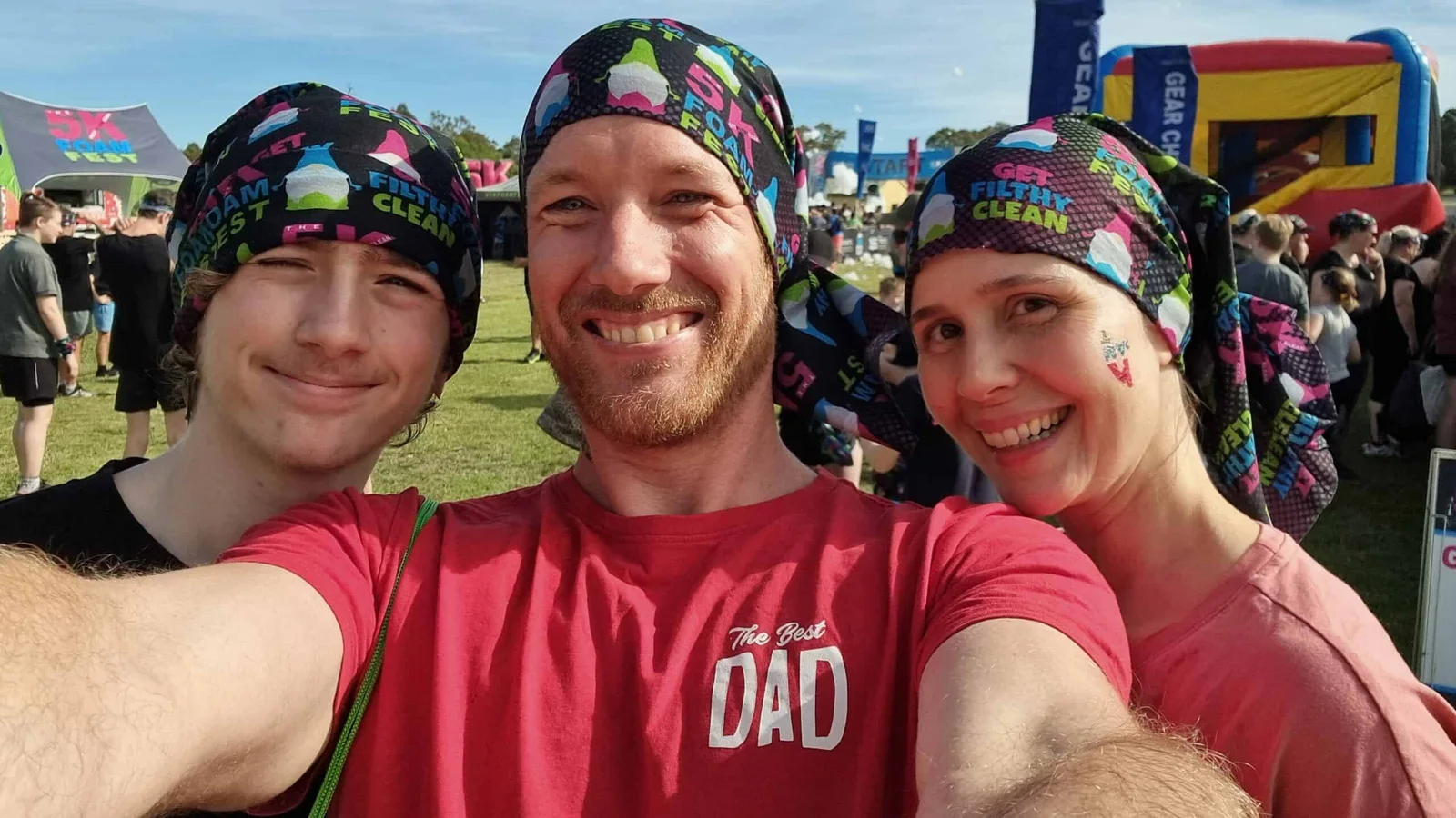Henk in a 'The Best Dad' shirt taking a selfie with his two children, all three wearing Foam Fest bandanas at an outdoor fun run