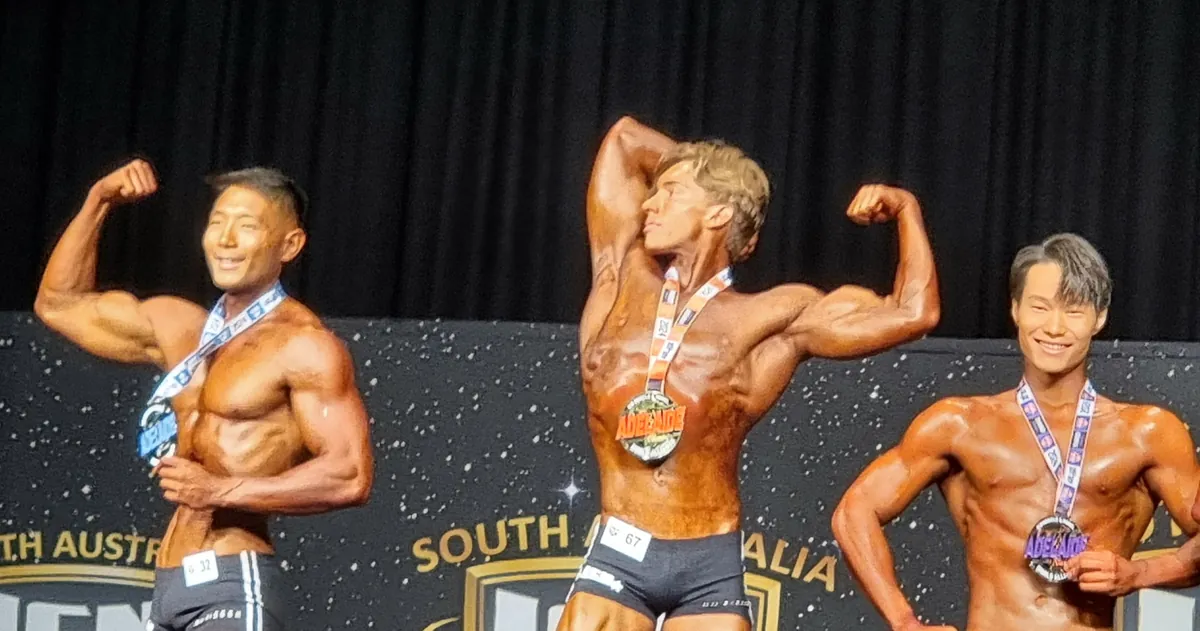 Mason on stage at a bodybuilding competition, holding a front double biceps pose with a medal around his neck